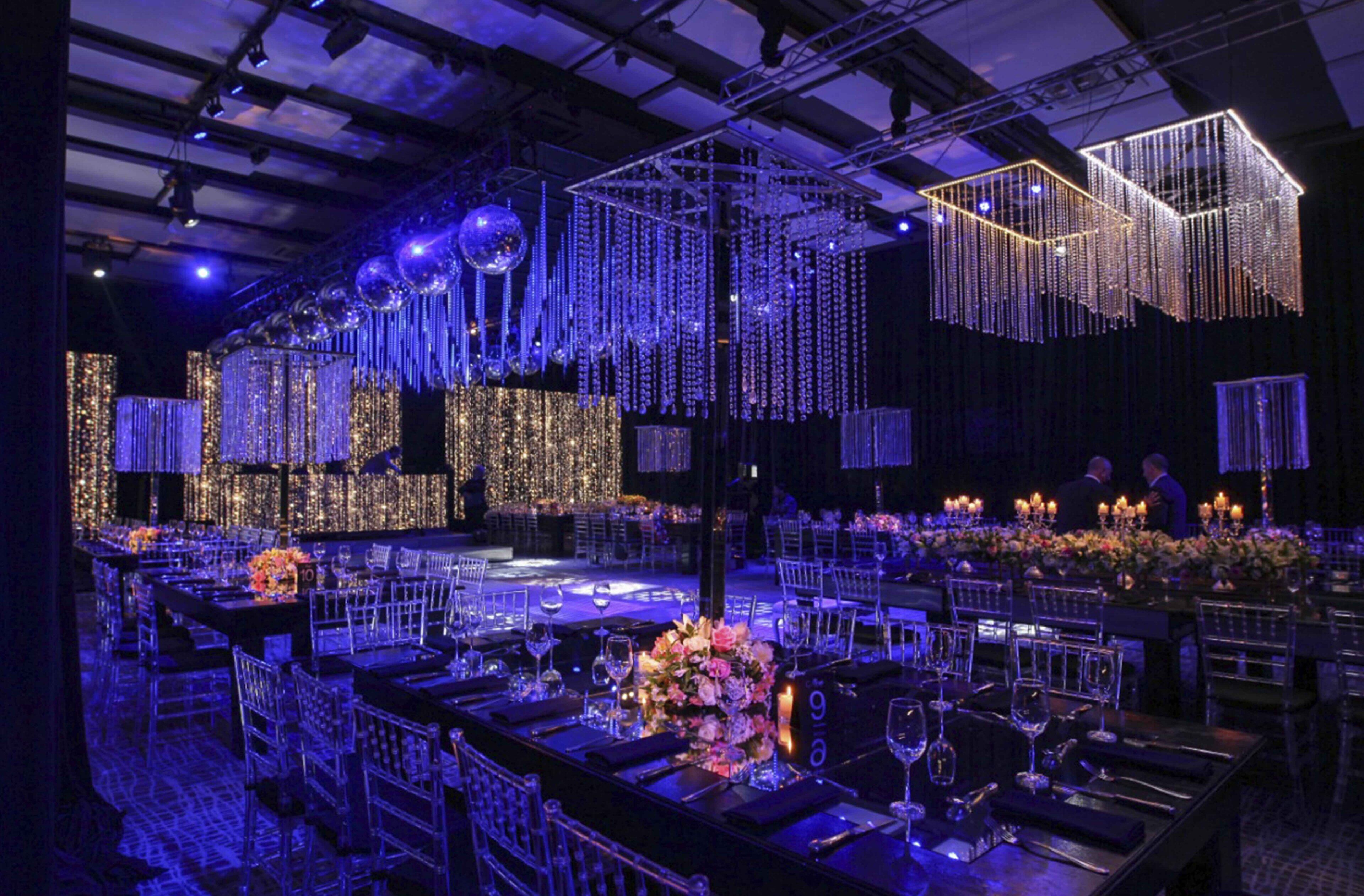 Un salón de banquetes con mesas dispuestas para un evento formal, que cuenta con un escenario con fondo de luces colgantes y una araña.