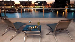 A terrace with two chairs and a table, overlooking a waterfront with boats and buildings in the background.