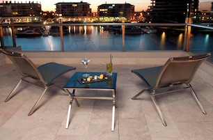 Terraço com duas cadeiras e uma mesa, com vista para a orla marítima com barcos e edifícios ao fundo.