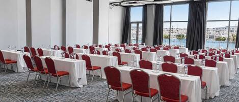 A conference room with rows of tables and chairs, large windows with a city view, and a carpeted floor.