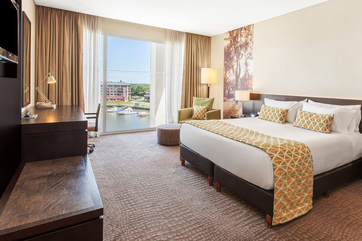 Habitación de hotel con una cama grande, un escritorio, una silla, un pequeño reposapiés y vistas al puerto deportivo.