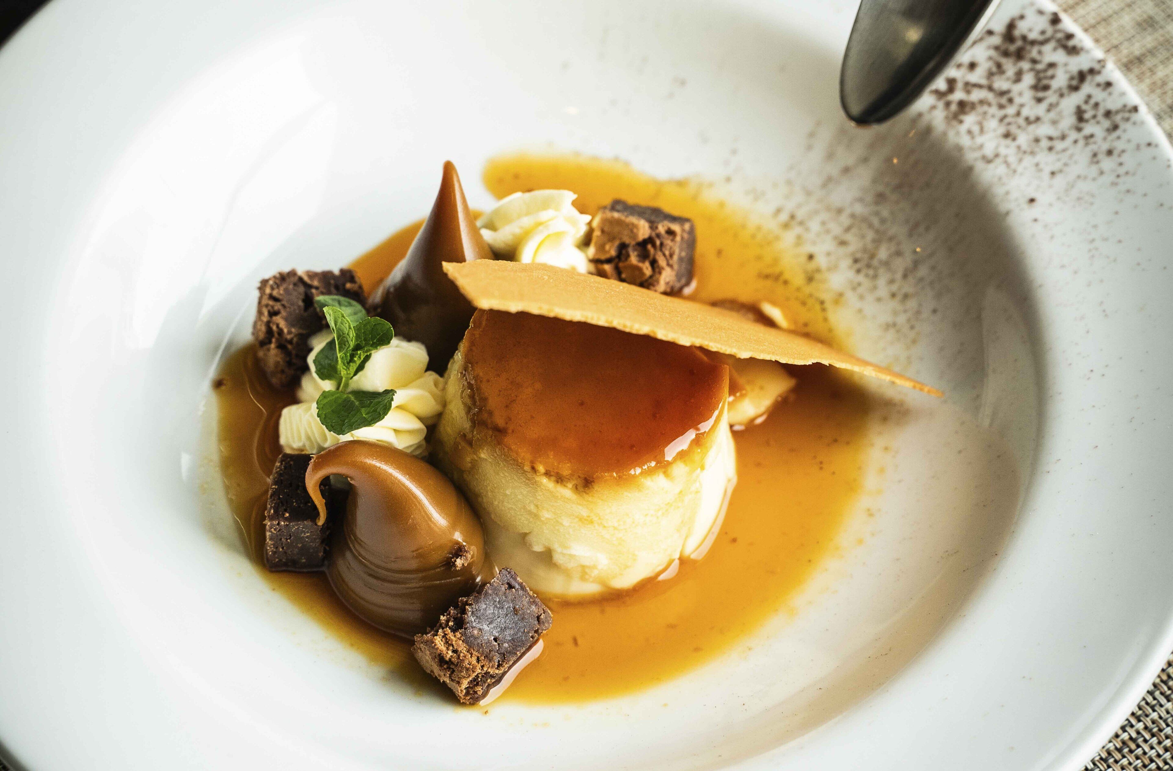 A dessert plate with a caramel flan, chocolate pieces, and a mint leaf garnish.