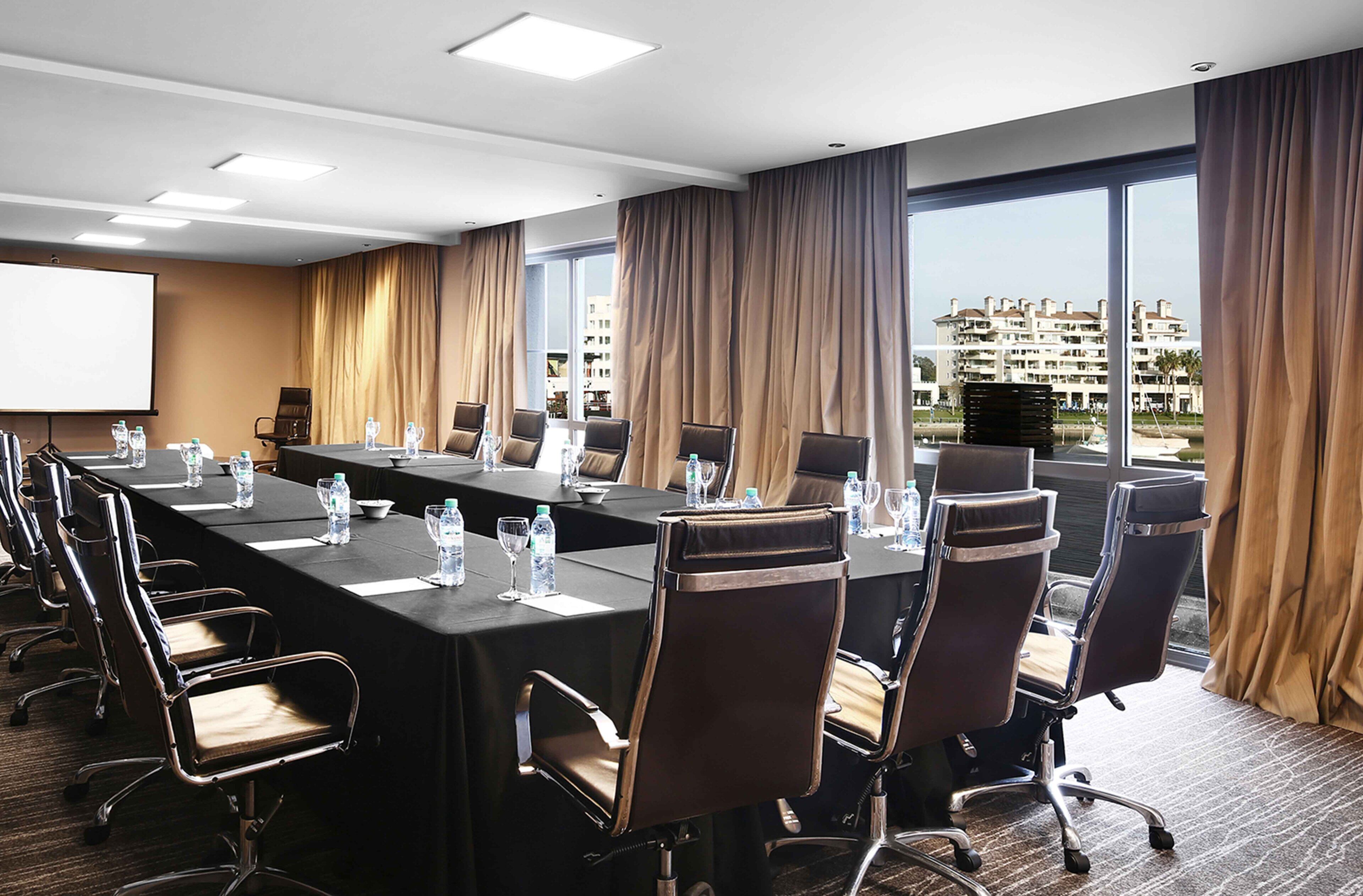 A conference room with a long table, chairs, a projector screen, and a view of buildings through large windows.