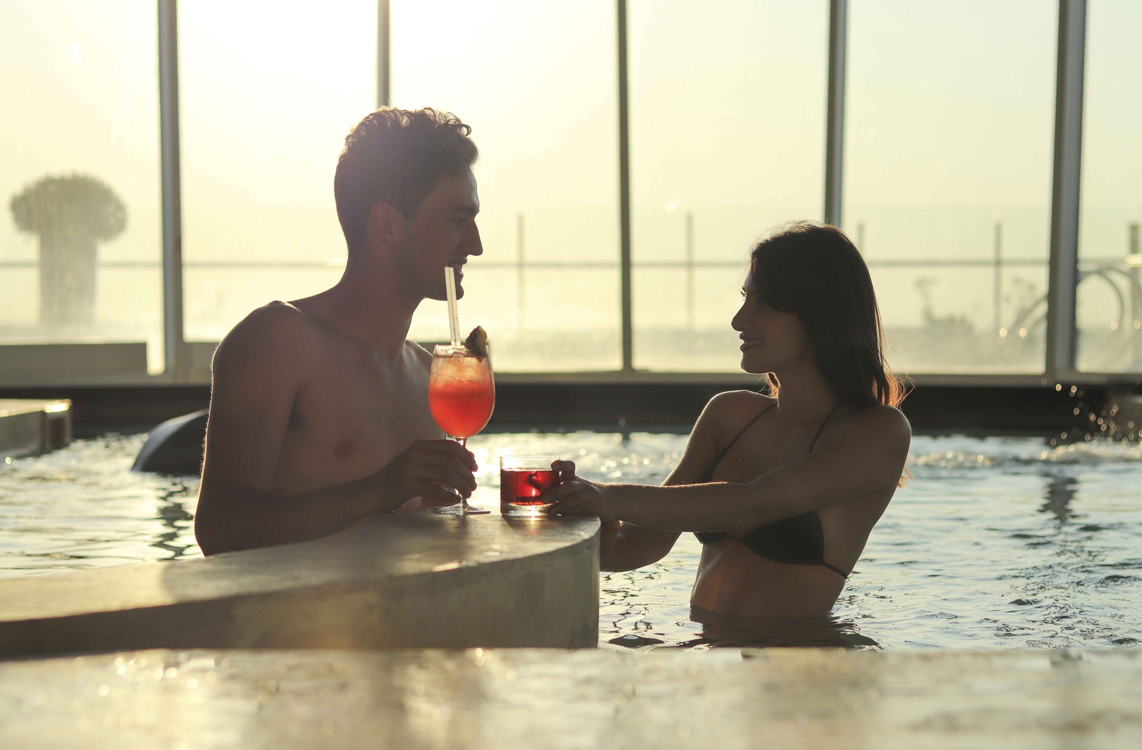 A couple in a heated pool with a drink, large windows, and a view of the water.