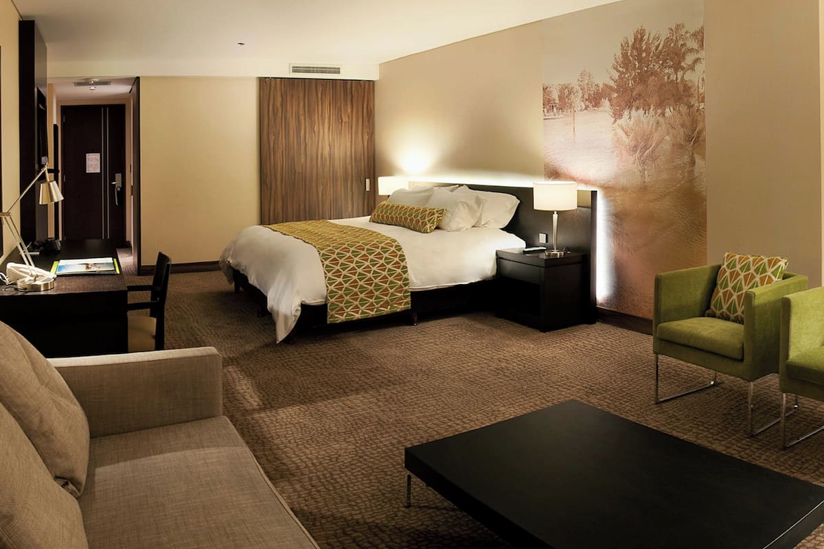 Una habitación de hotel con una cama, mesitas de noche, un escritorio, un sillón verde y una mesa de centro.