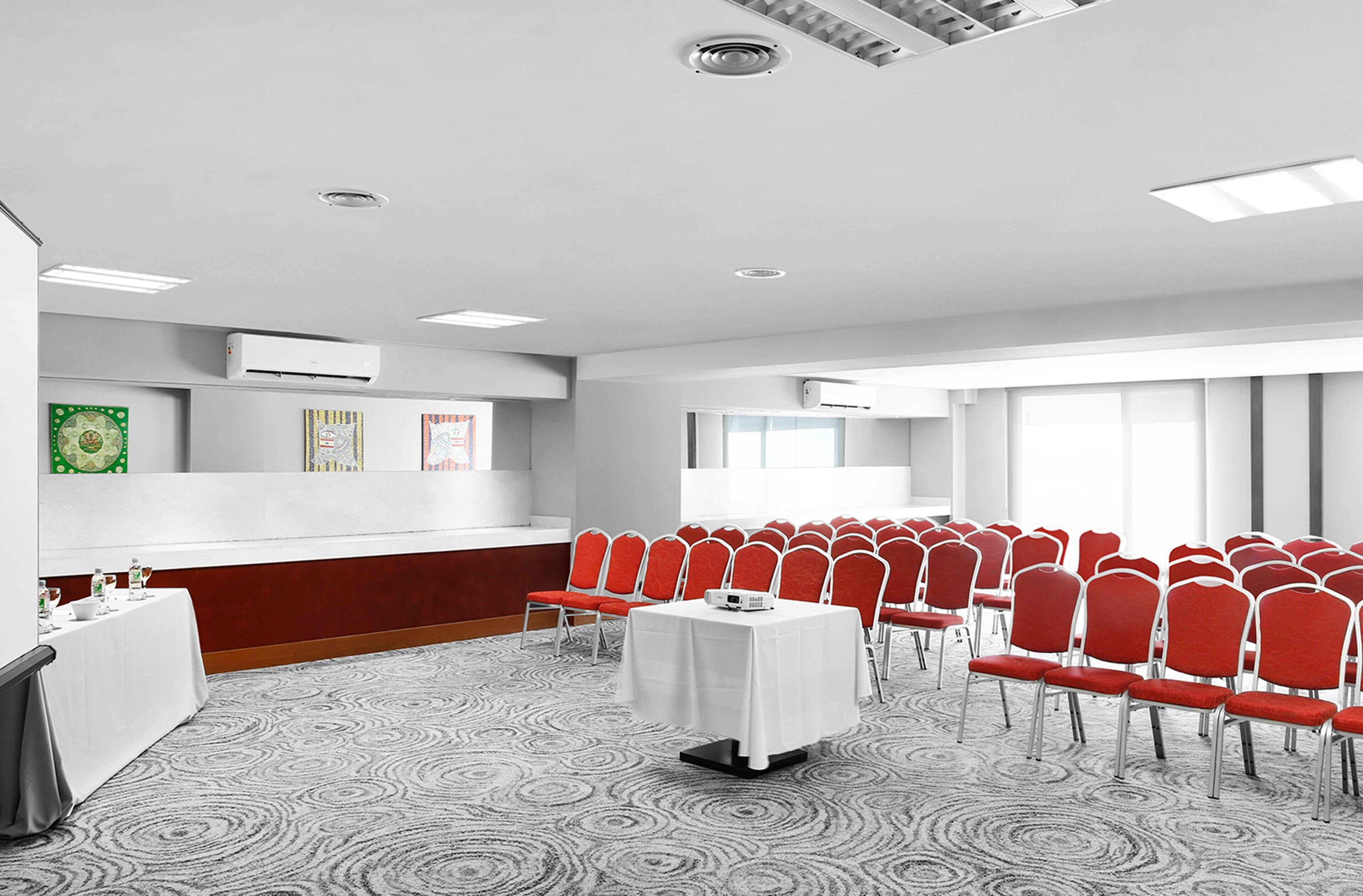 A conference room with rows of red chairs, a white table with a projector, and a patterned carpet.