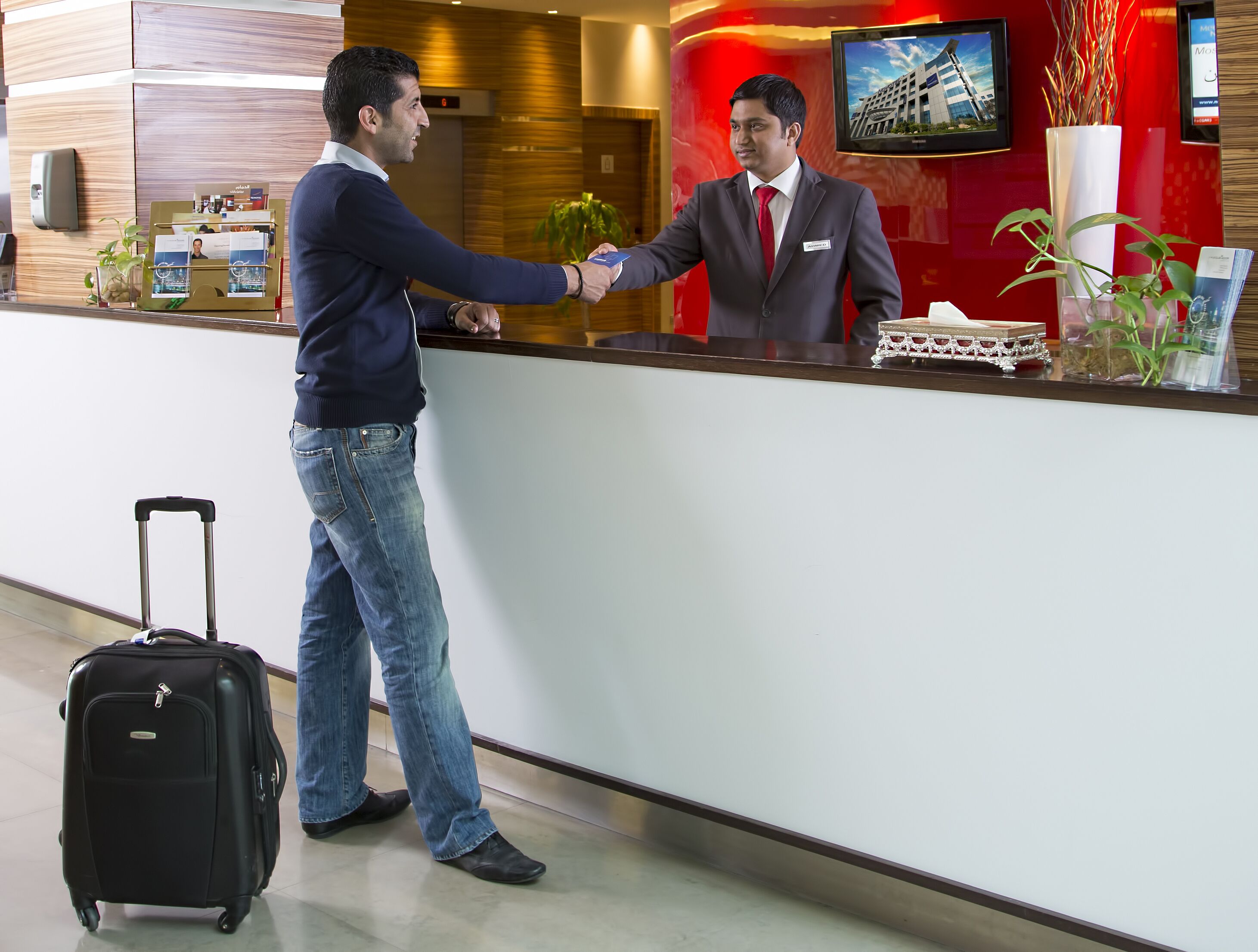 Ein Hotel-Empfangsbereich mit einem Holzbüro, einem digitalen Bildschirm, der ein Gebäude zeigt, und einer kleinen Pflanze auf der Theke.