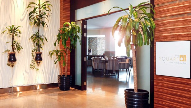 A modern lobby with potted plants, a reception desk, and a dining area visible through a glass partition.