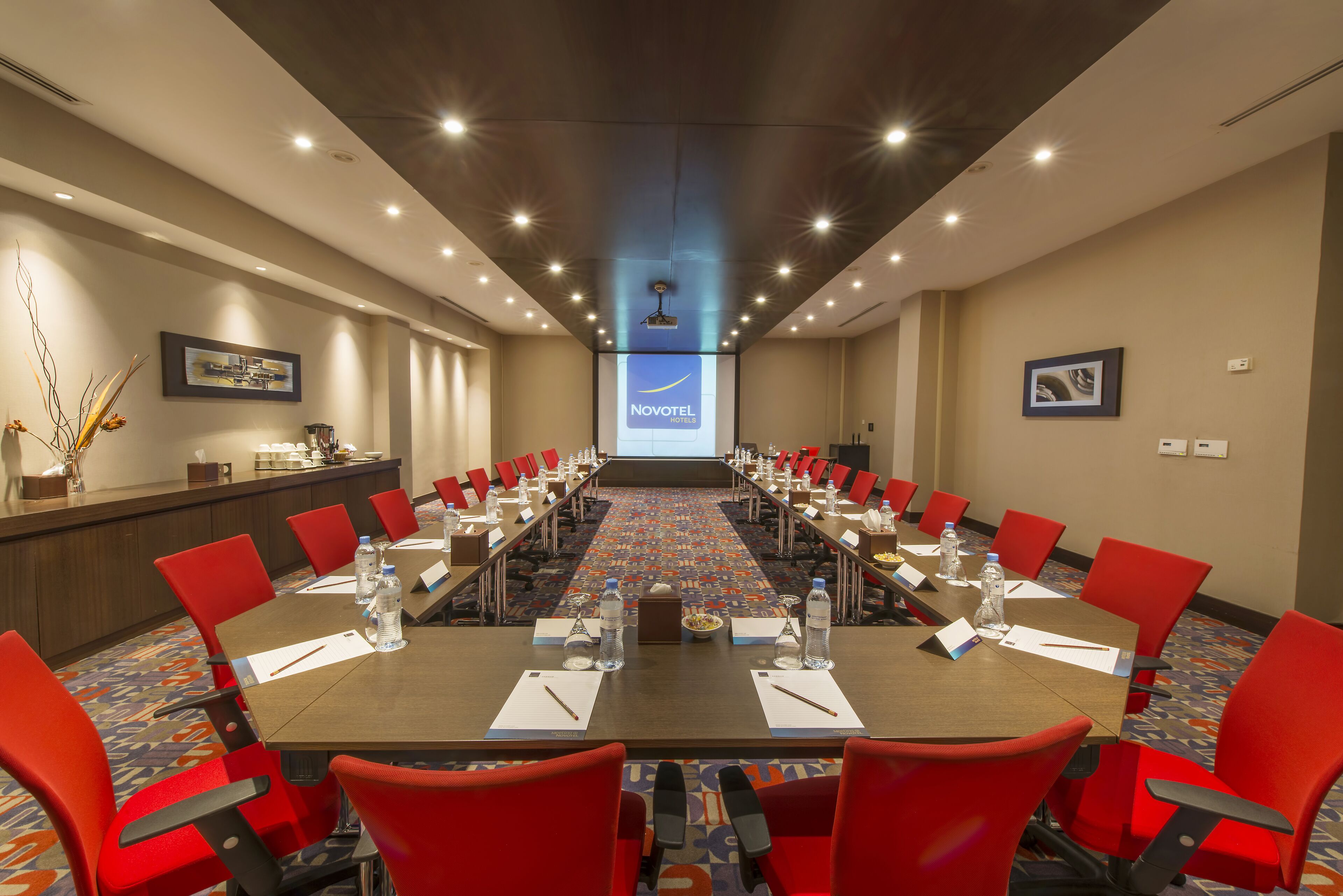 A conference room with a long table, red chairs, and a projector screen displaying the Novotel logo.