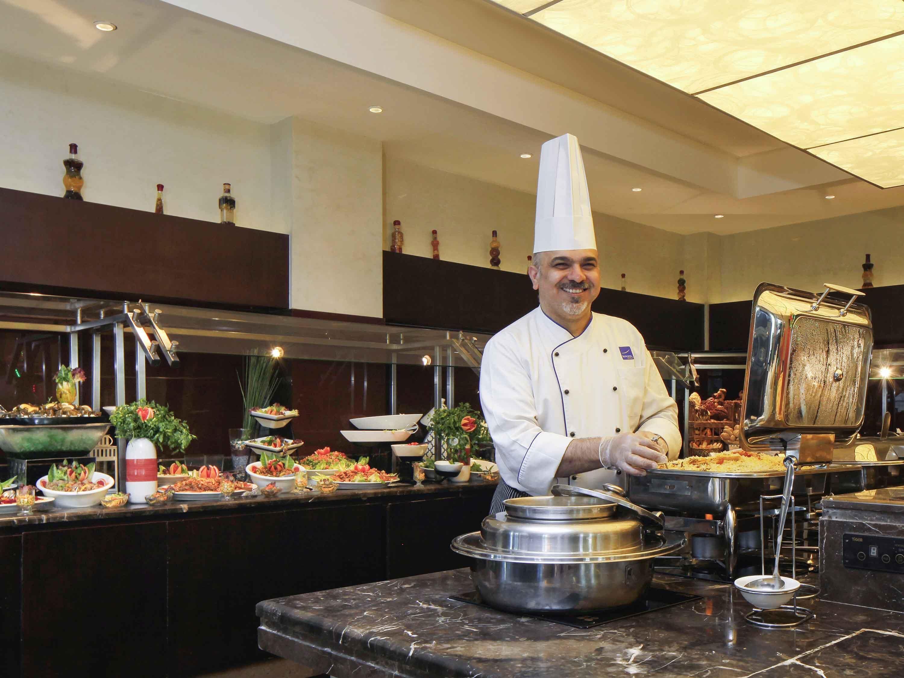 A chef in a white uniform with a tall hat, standing in a kitchen with a buffet setup.