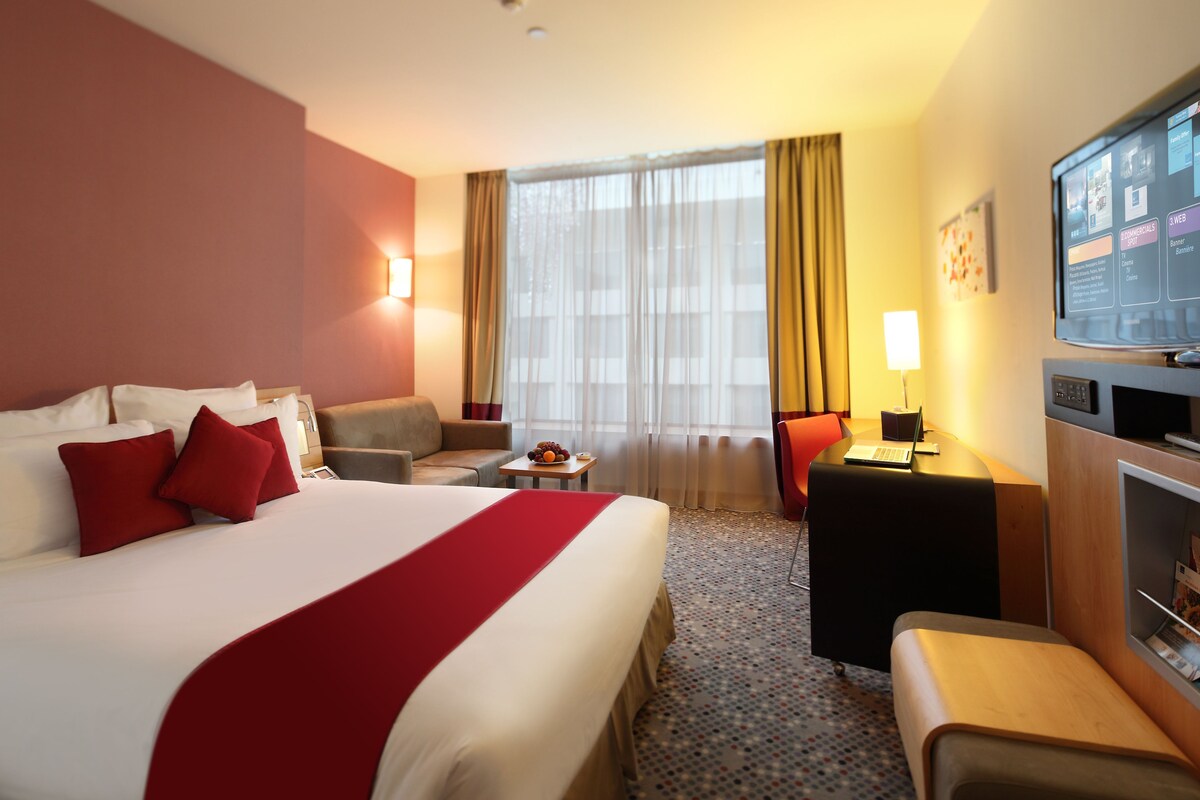 Une chambre d’hôtel avec un grand lit, un bureau, une chaise, une petite table avec un panier de fruits, une télévision et une fenêtre avec des rideaux.
