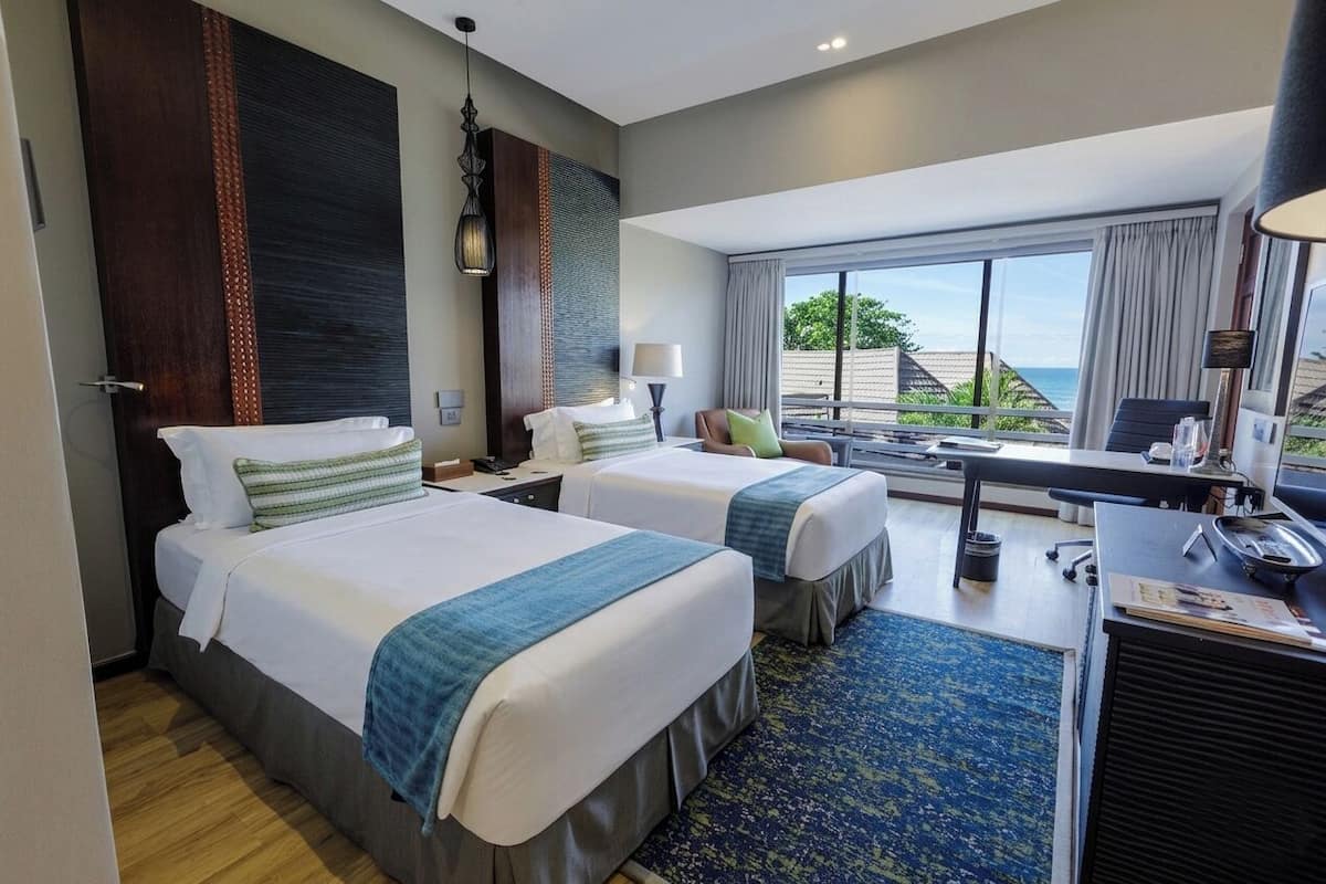 Une chambre d’hôtel avec deux lits, un bureau, une chaise, une lampe et une vue sur l’océan.