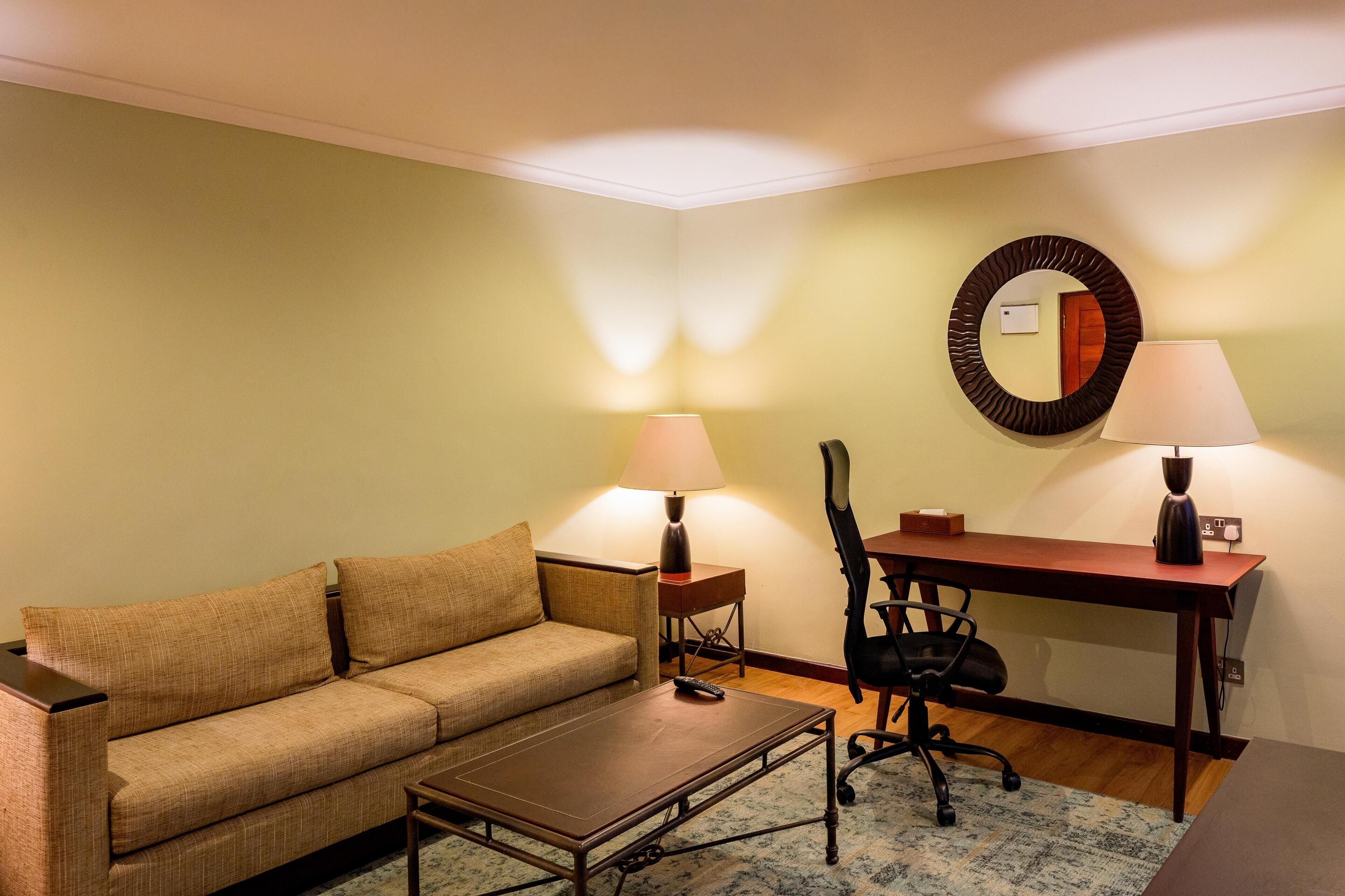 A furnished room with a sofa, a small table, a desk with a chair, two lamps, and a mirror on the wall.