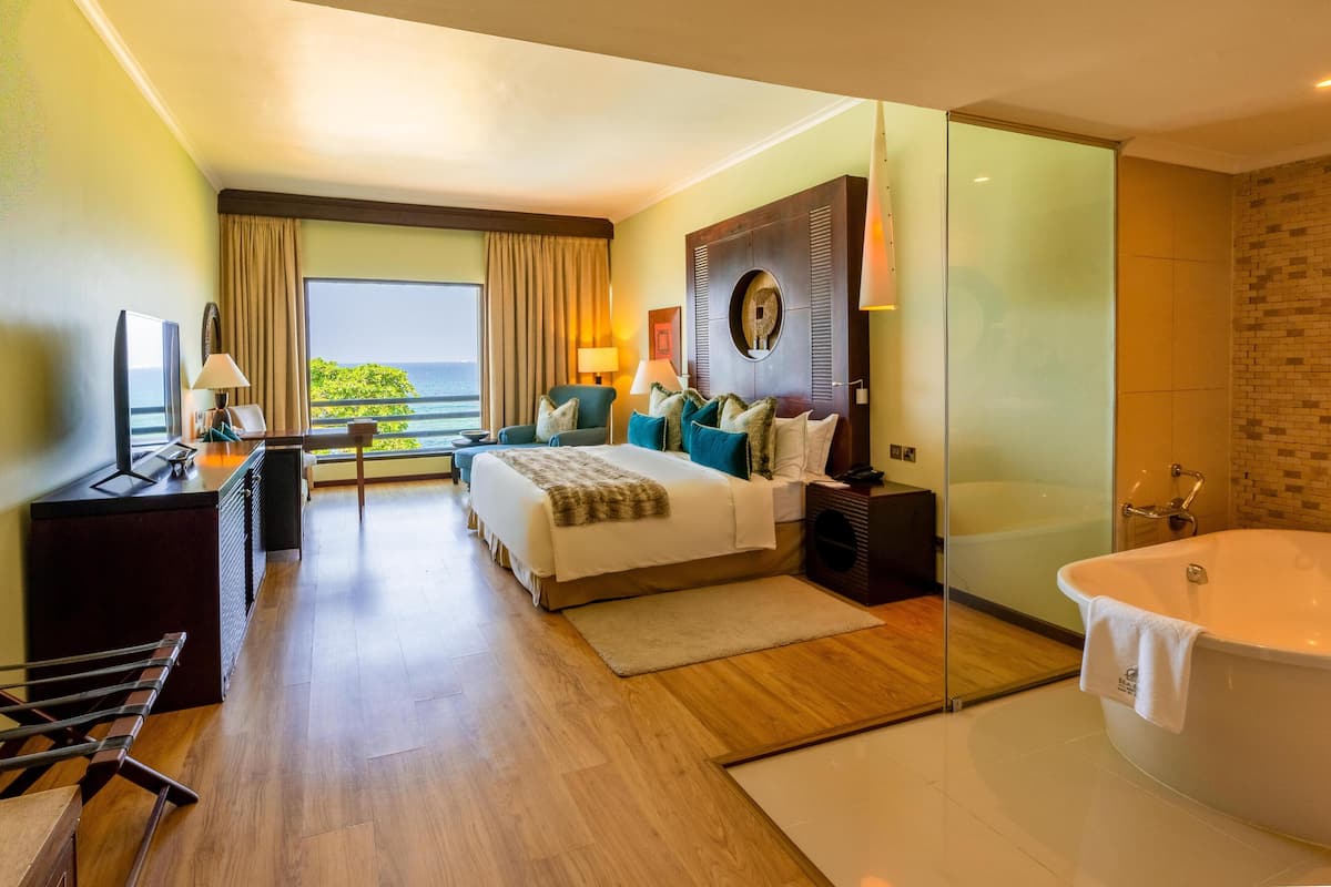 Une chambre d’hôtel comprenant un lit, un bureau, une chaise, une grande fenêtre avec vue, une salle de bain fermée par des parois de verre et dotée d’une baignoire, ainsi qu’un petit balcon.