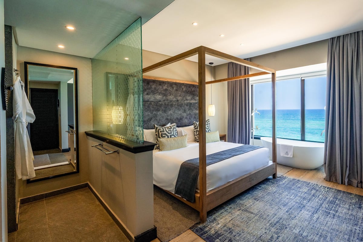 Une chambre d’hôtel moderne dotée d’un lit à baldaquin, d’une douche en verre, d’une grande fenêtre donnant sur la mer et d’une salle de bain séparée.