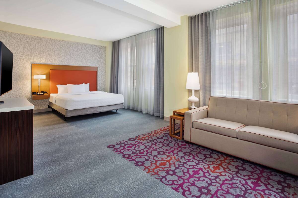 A hotel room with a bed, a sofa, a desk with a lamp, and a TV.
