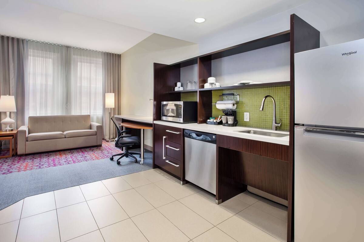 A compact hotel kitchen with a sofa, a desk, a dishwasher, a microwave, a refrigerator, and a sink.