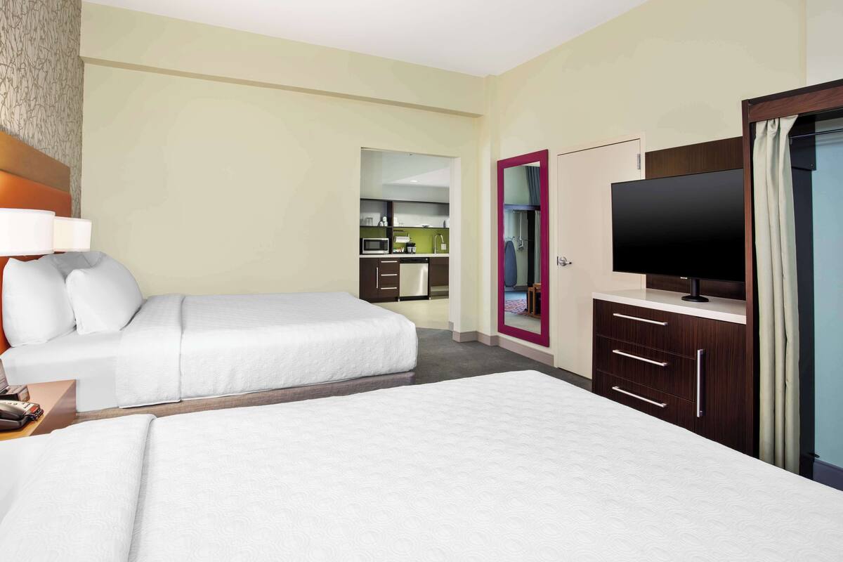 A hotel room with two beds, a TV, a wardrobe, and a kitchenette in the background.