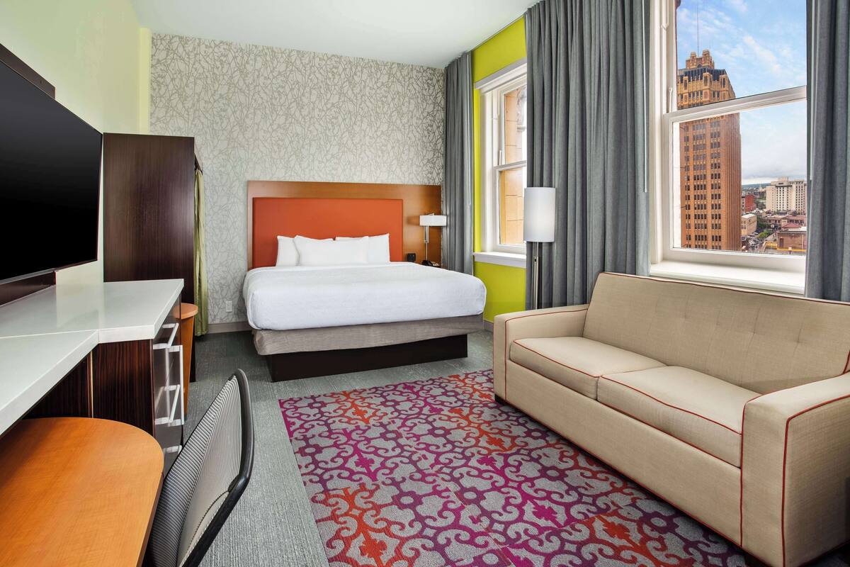 A hotel room with a bed, a sofa, a desk, a TV, and a window with a city view.