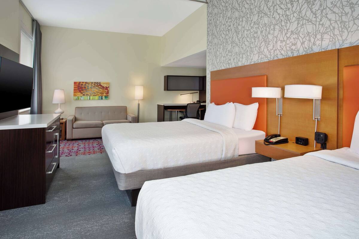 A hotel room with two beds, a desk with a lamp, a TV, and a painting on the wall.