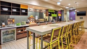 A modern cafe with a long communal table, yellow chairs, and a variety of food and drink options. - Home2 Suites by Hilton San Antonio Downtown - Riverwalk, TX (San Antonio)