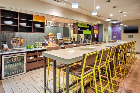A modern cafe with a long communal table, yellow chairs, and a variety of food and drink options.