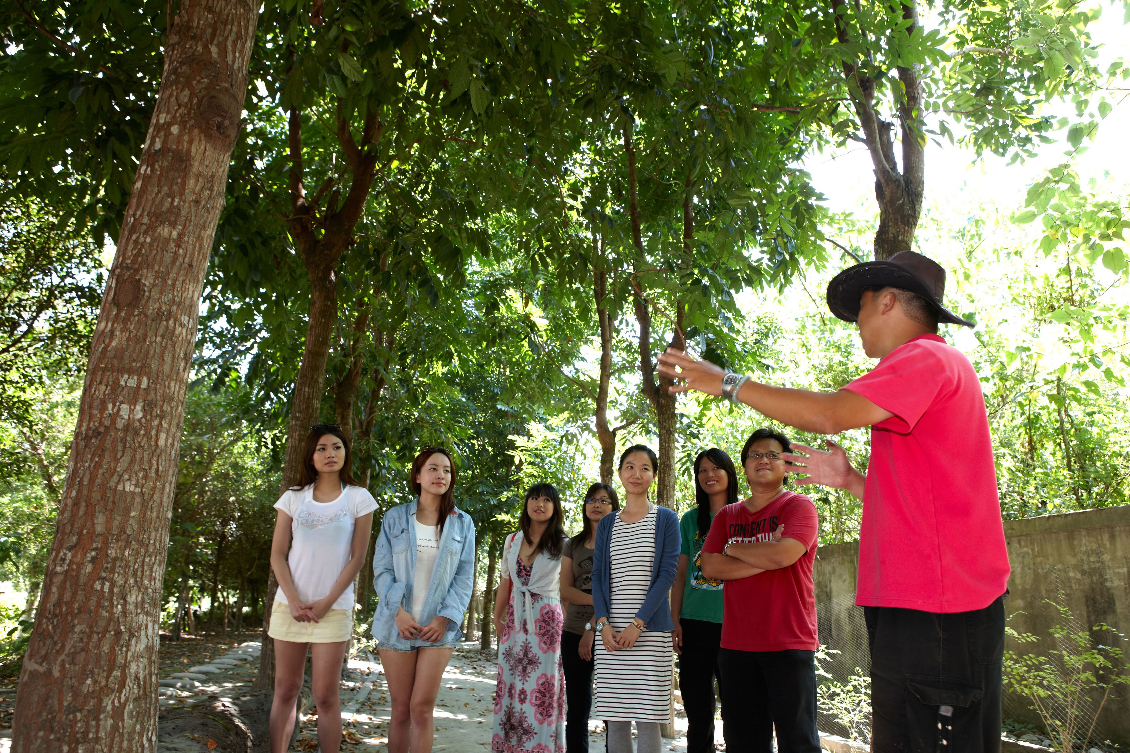 Un grupo de personas de pie al aire libre, quizás en un parque o jardín, con árboles y un sendero al fondo.