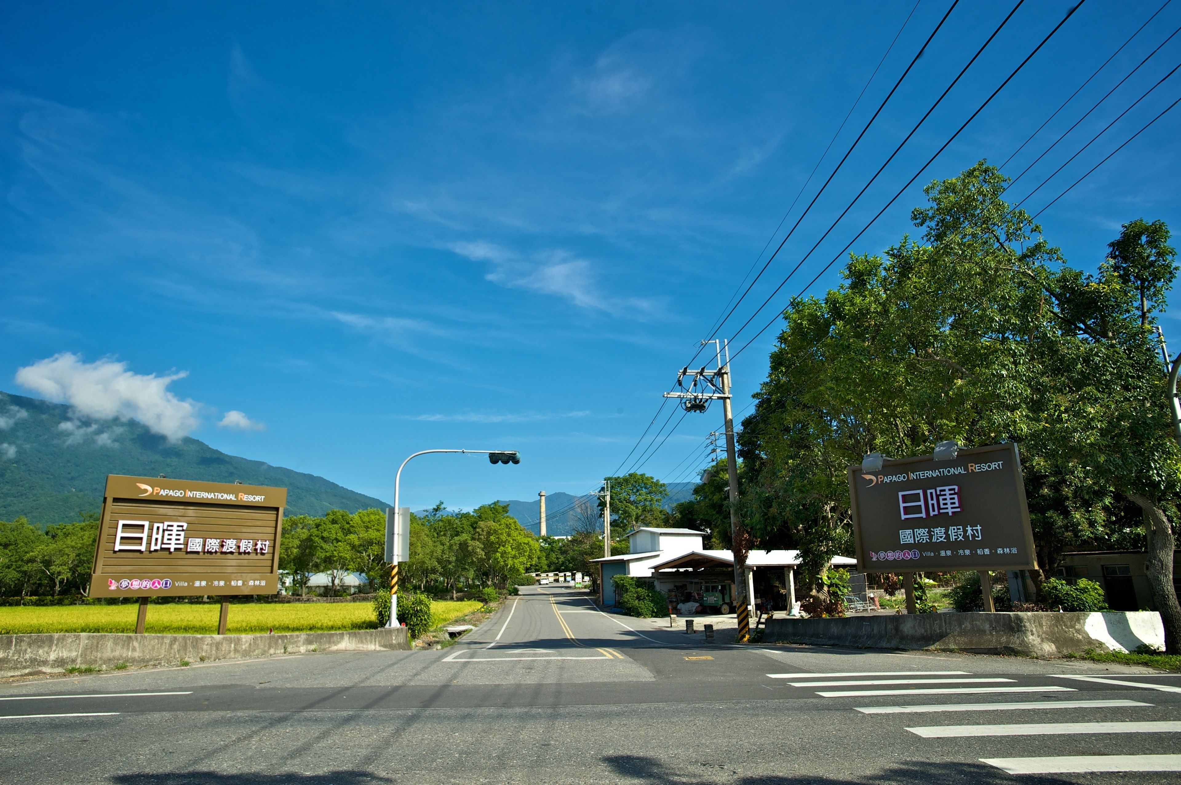 A road leading to a resort with bilingual signage, a clear sky, and lush greenery.