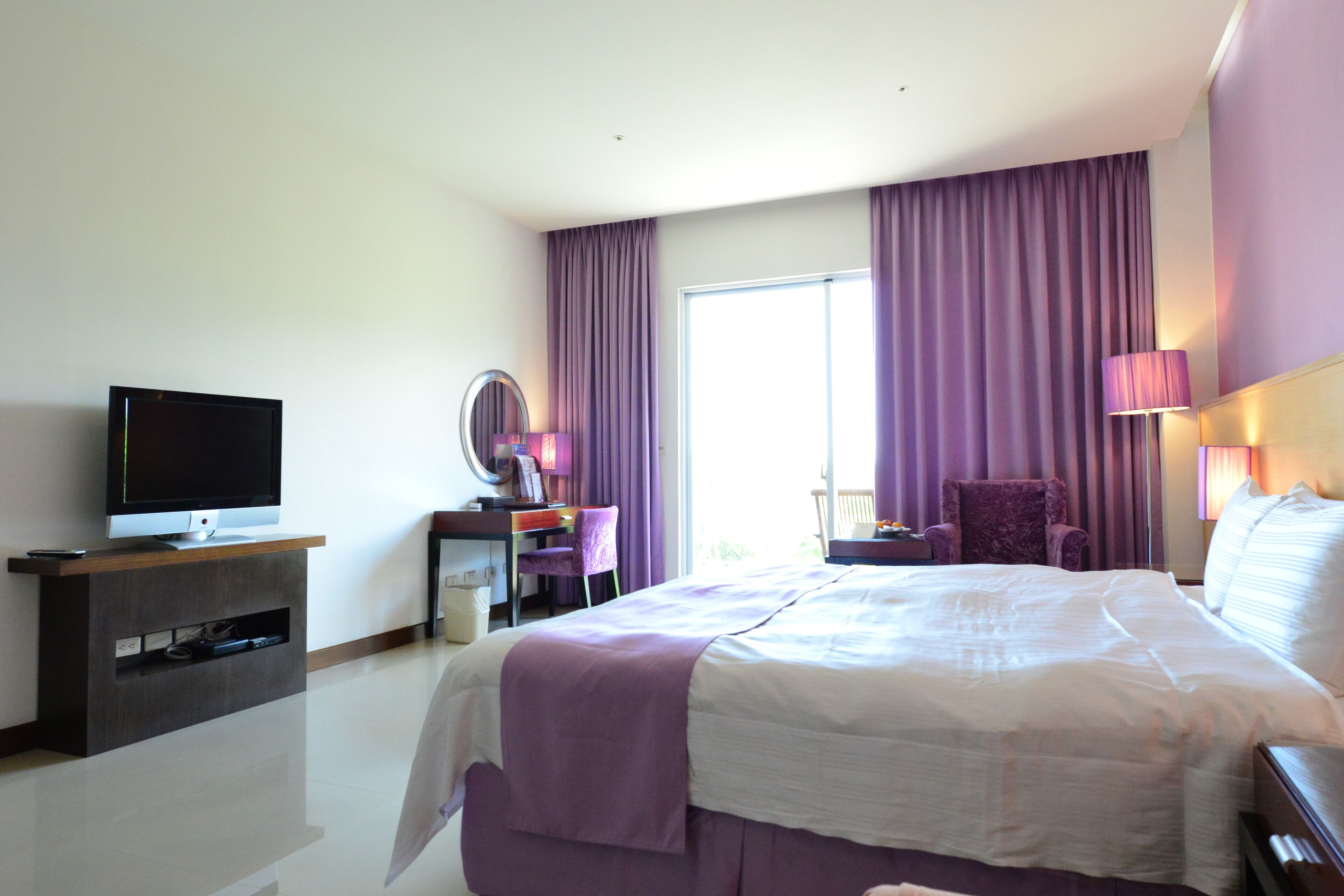 A hotel room with a large bed, a television on a wooden stand, a desk with a chair, and a window with purple curtains.