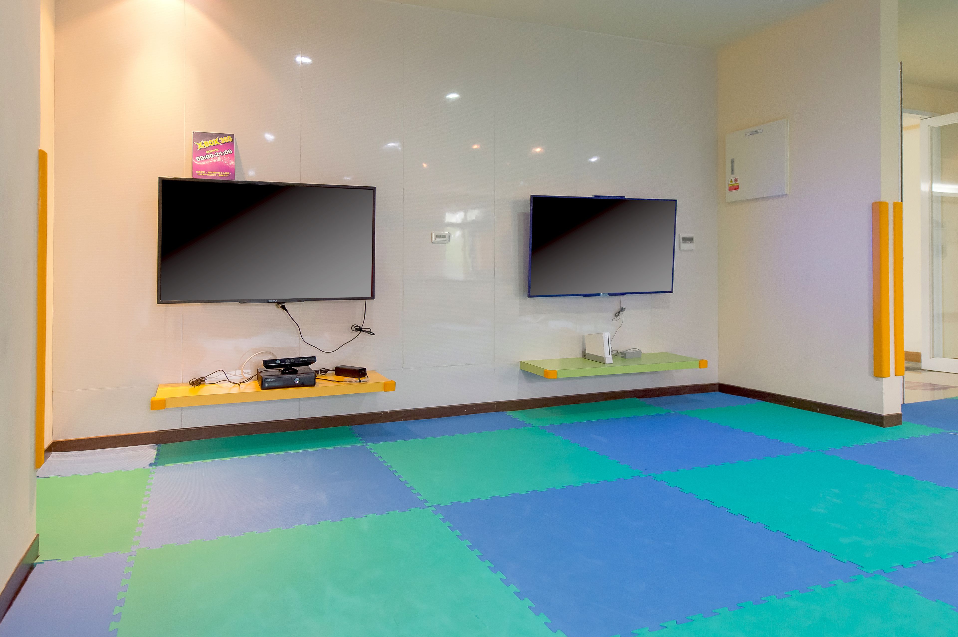 A room with two flat-screen TVs mounted on the wall, a small desk with a computer, and a colorful interlocking floor mat.