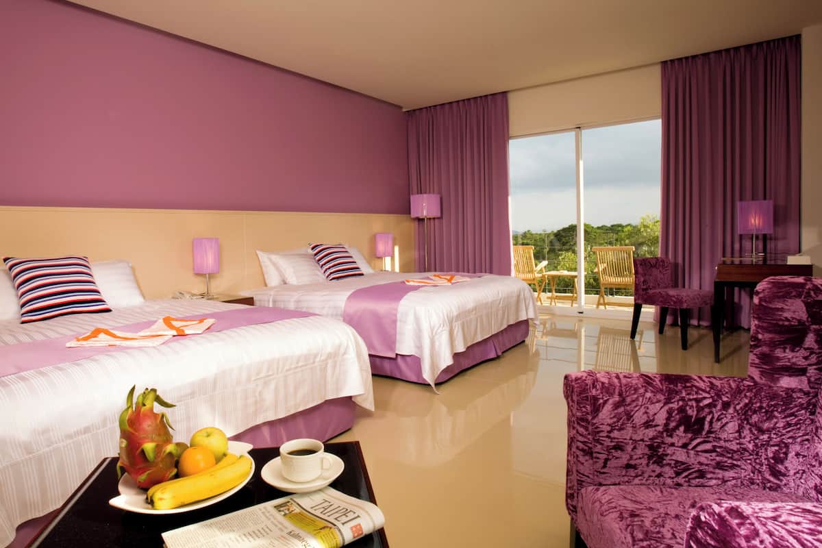 Habitación de hotel con dos camas, zona de comedor con mesa y sillas, y balcón con vistas.