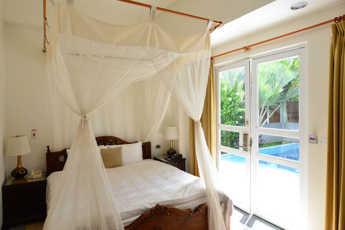 Un dormitorio con cama con dosel, una mesita de noche con lámpara y vista a una piscina a través de un ventanal.