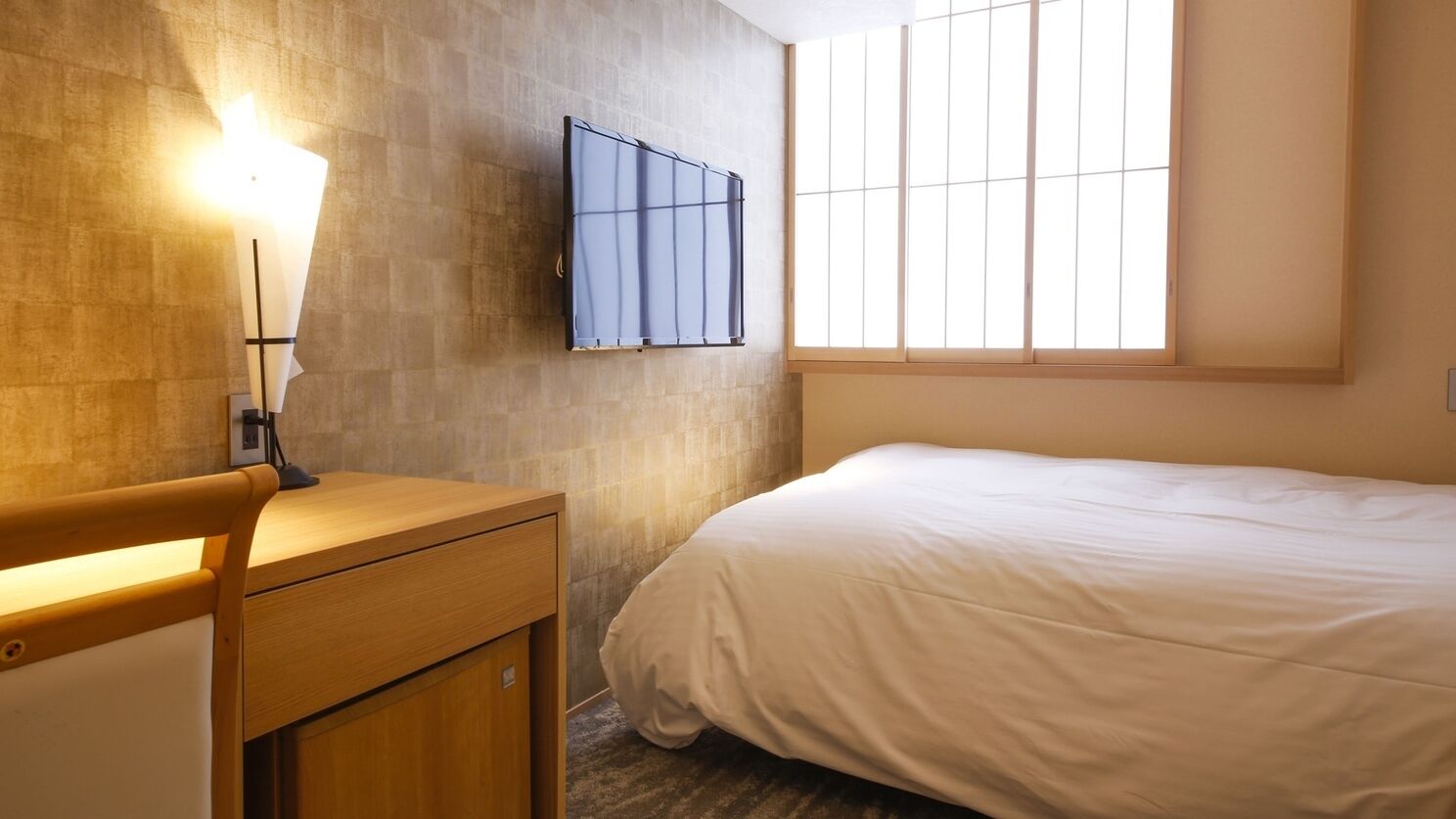 Et hotelværelse med seng, skrivebord med lampe, et vindue med rullegardiner og et fjernsyn monteret på væggen.