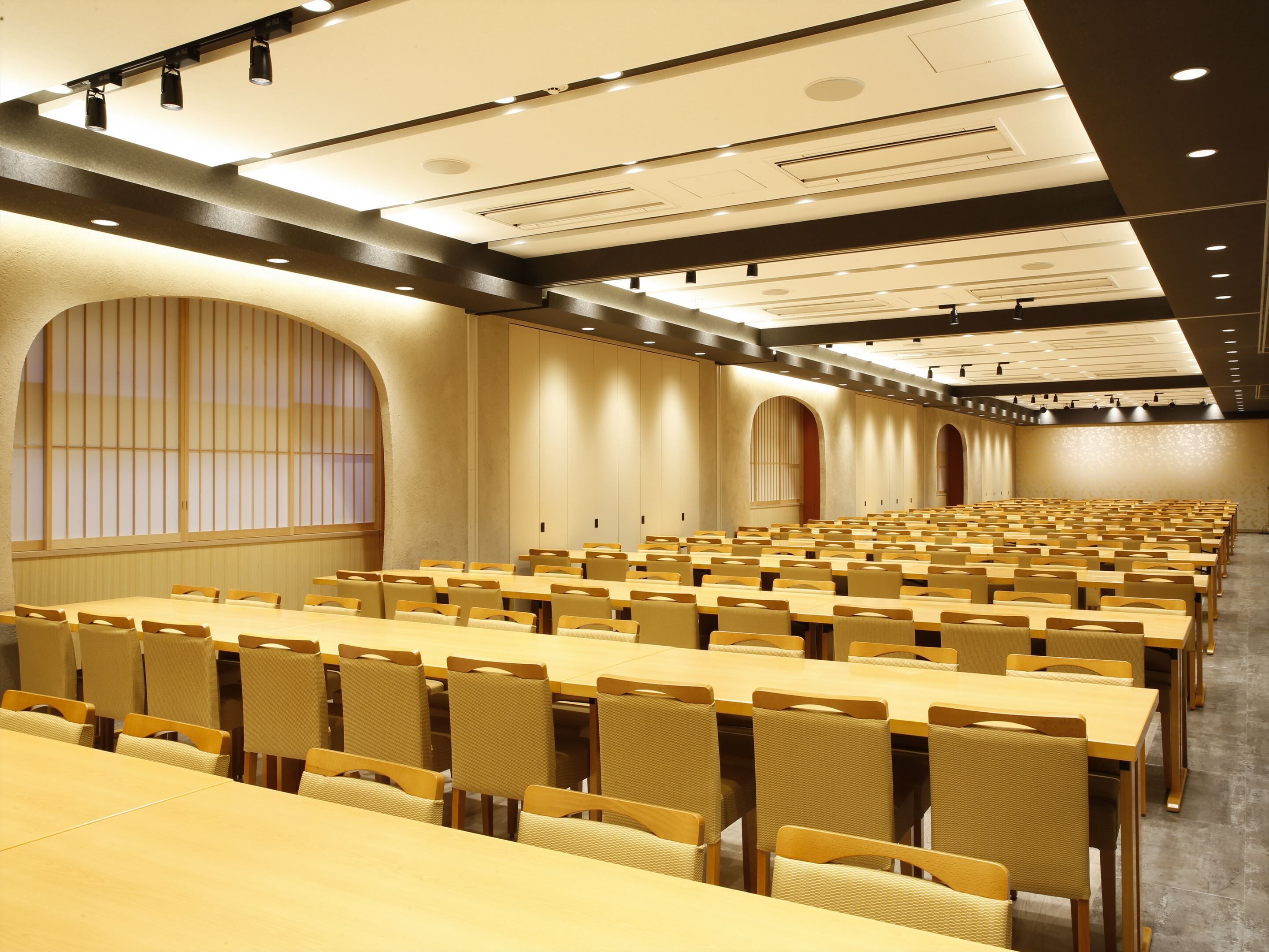 A conference room with rows of tables and chairs, large windows with vertical blinds, and a high ceiling with recessed lighting.