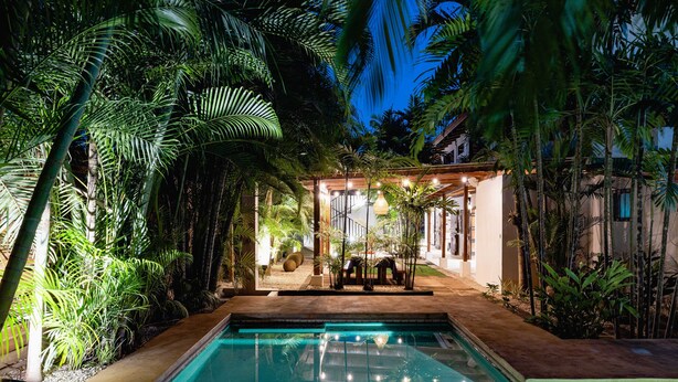A swimming pool surrounded by lush tropical plants, with a covered seating area and a building in the background.