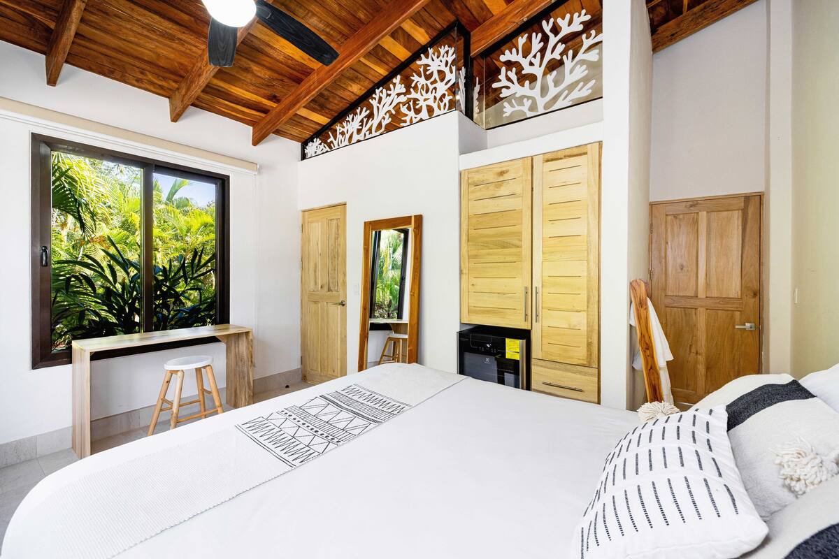 Ein Schlafzimmer mit einem großen Bett, Holzboden, einem Schreibtisch mit Hocker und Blick ins Grüne durch ein Fenster.