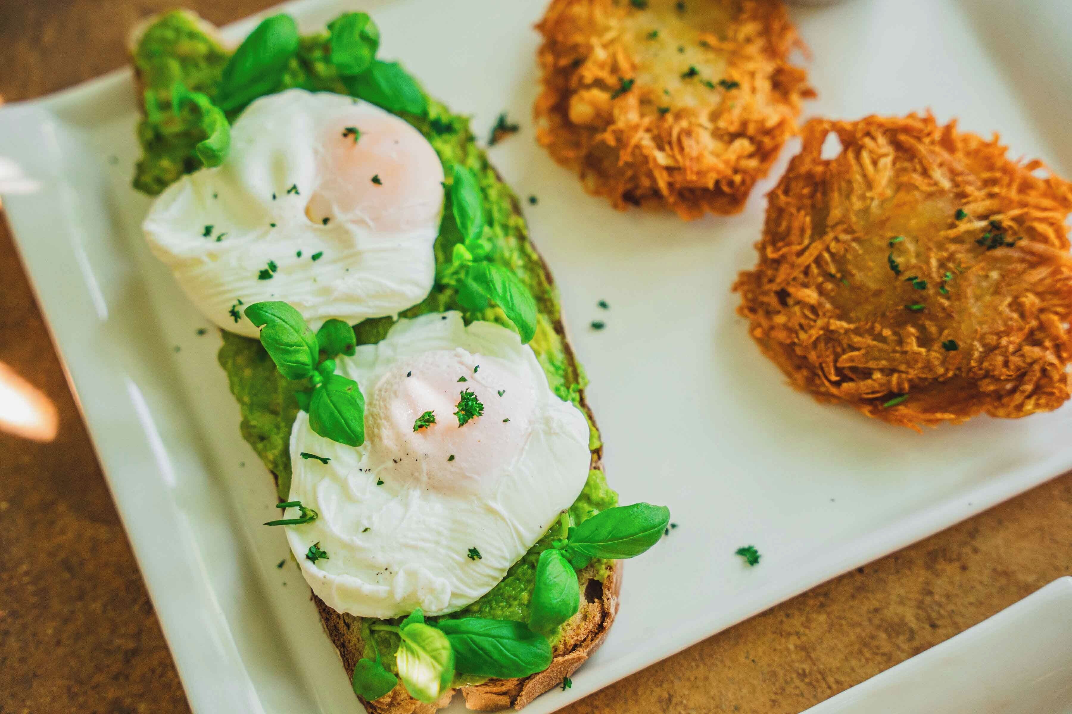 Un plato con un sándwich cubierto de aguacate, huevos pochados y albahaca, junto a dos tortitas de patata crujientes.