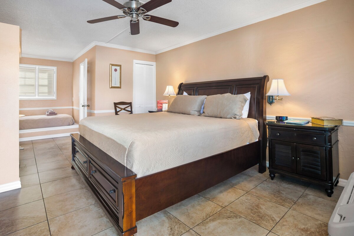 A bedroom with a large bed, a nightstand, a ceiling fan, and a window with blinds.