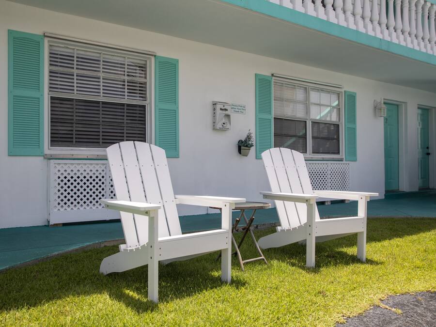 A white building with teal shutters, two white Adirondack chairs, and a small table.