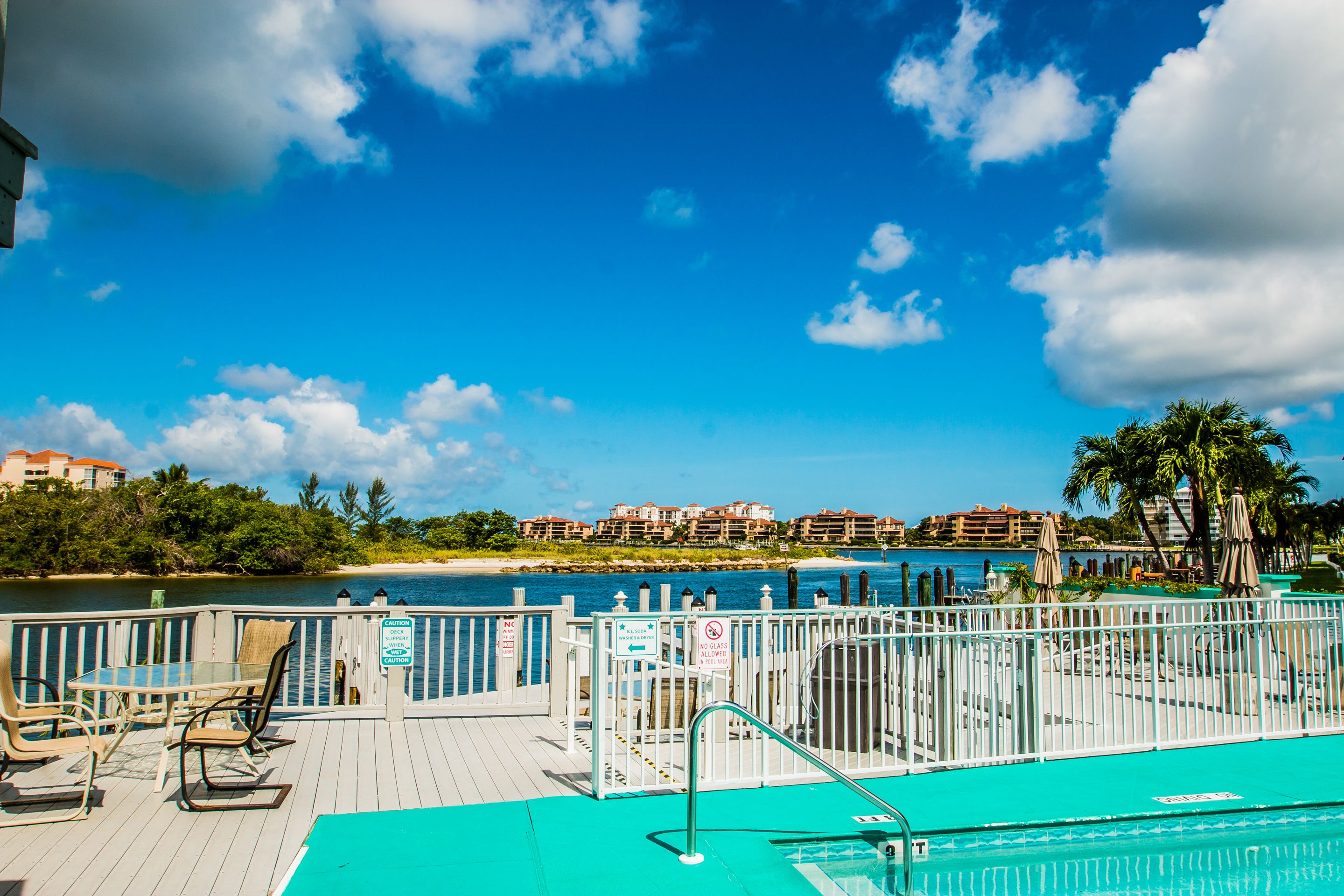 A deck with a pool, chairs, and a view of the beach and buildings.