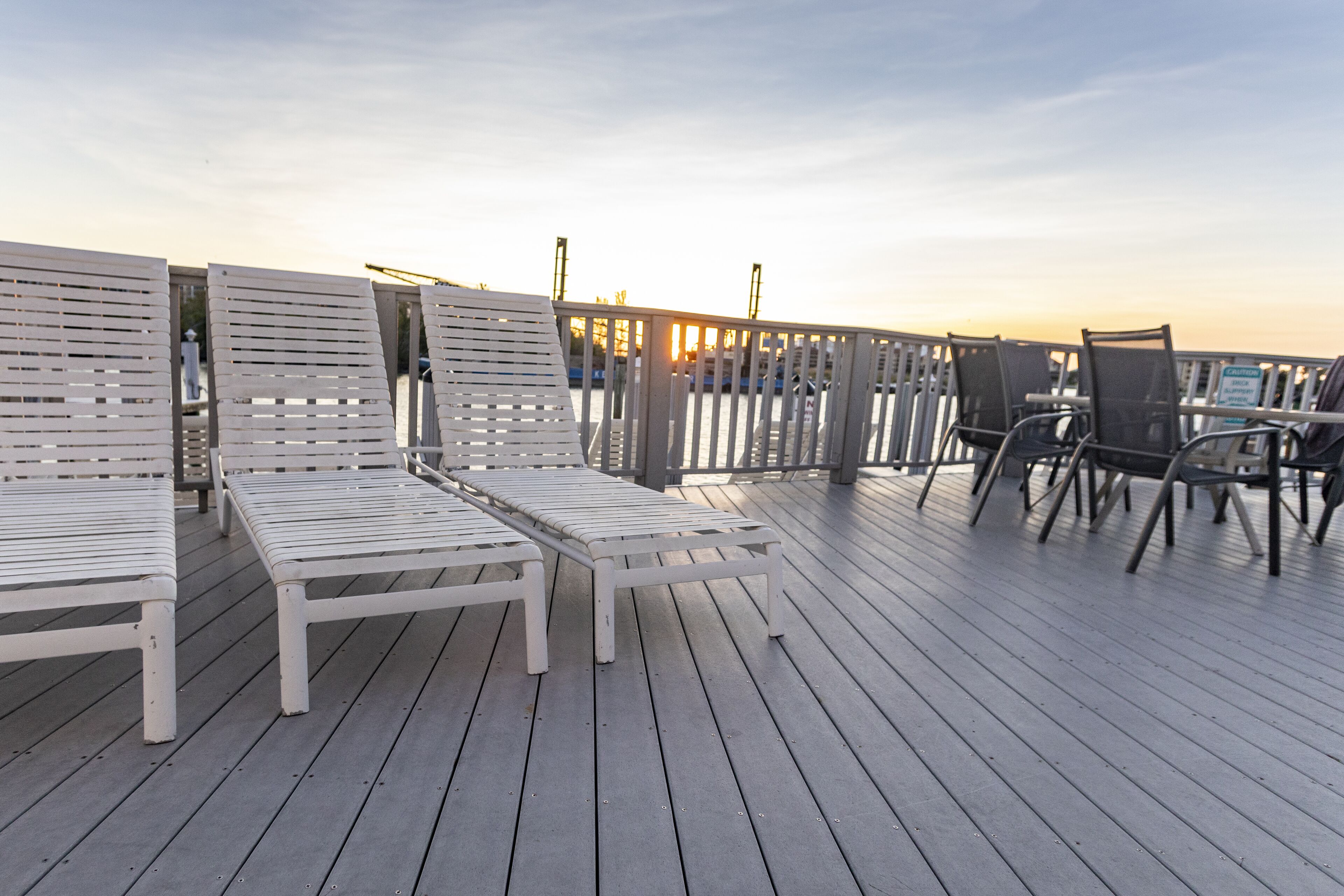 A wooden deck with white lounge chairs and metal chairs, overlooking a body of water.