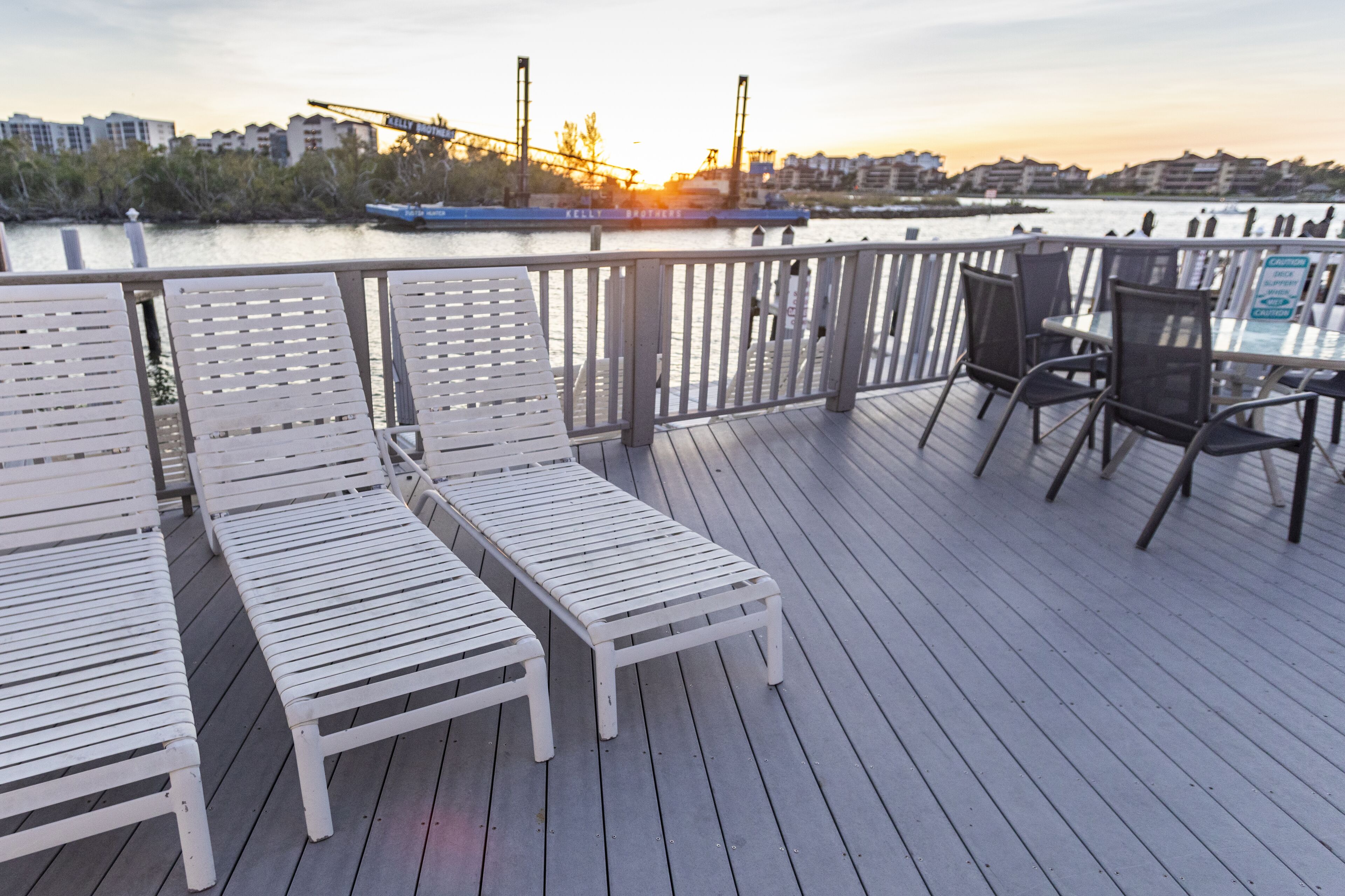 Een terras met witte ligstoelen en een tafel, met uitzicht op een waterpartij en een stadsachtergrond.