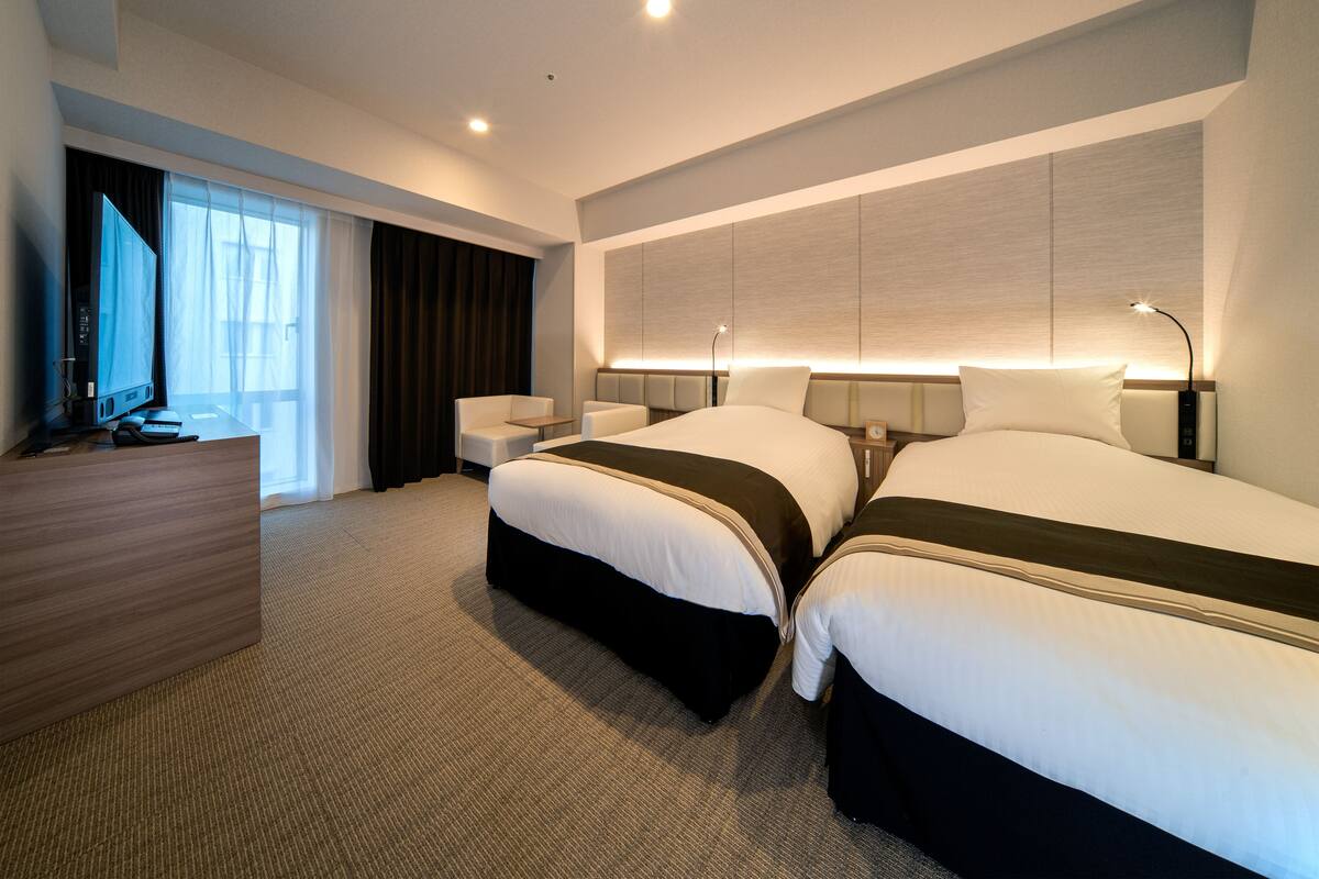 Une chambre d’hôtel avec deux lits, un bureau, une chaise et une grande fenêtre.