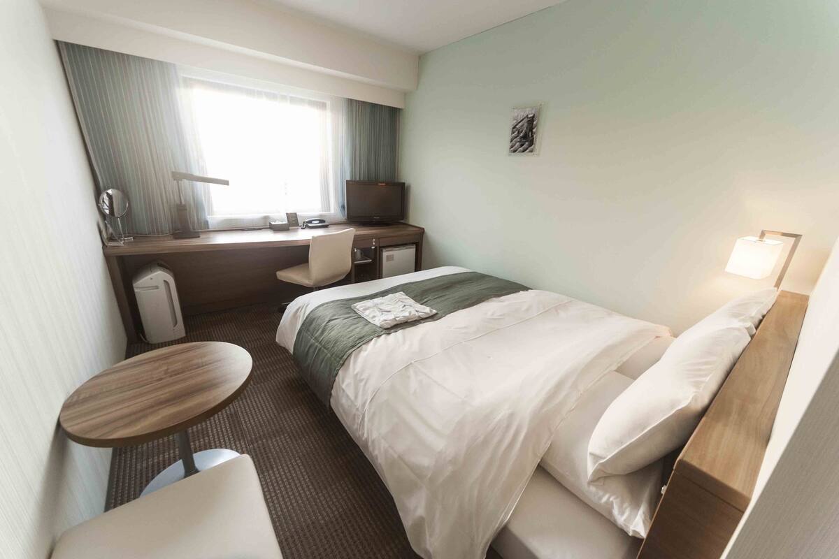 Une chambre d’hôtel comprenant un lit, un bureau, une chaise, une petite table, un téléviseur et une fenêtre avec des rideaux.
