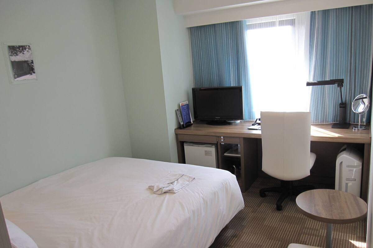 Une chambre d’hôtel comprenant un lit, un bureau avec un ordinateur, une chaise, un petit réfrigérateur et une fenêtre avec des rideaux.