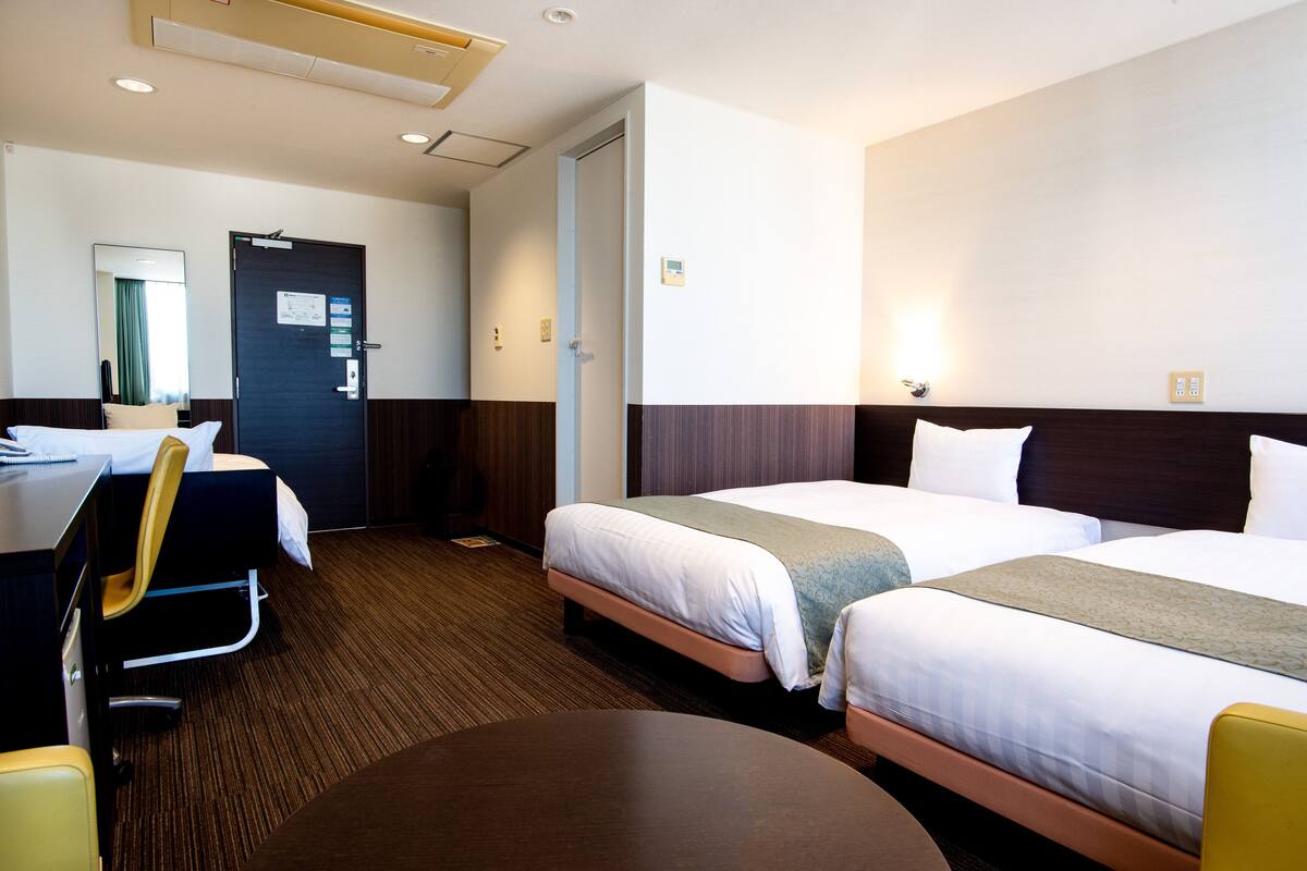 A hotel room with two beds, a desk with a chair, a round table, and a door.