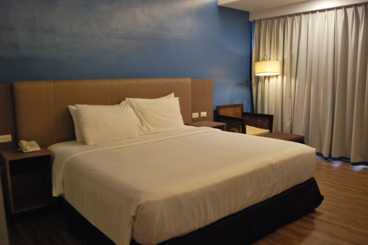 Habitación de hotel con una cama grande, una mesita de noche con un teléfono, una lámpara y una ventana con cortinas.