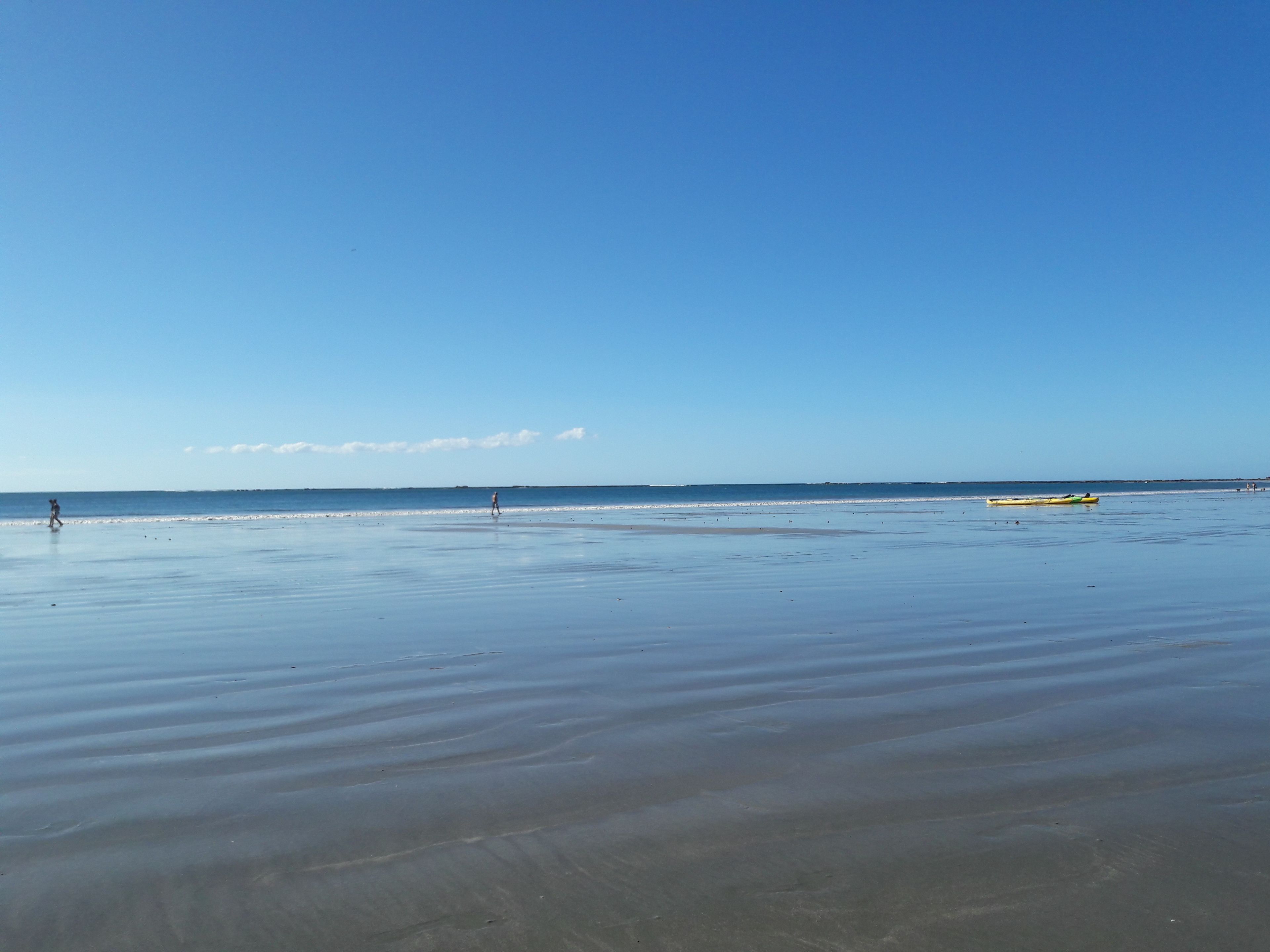 A beach with a calm sea, a clear sky, and a few people walking.