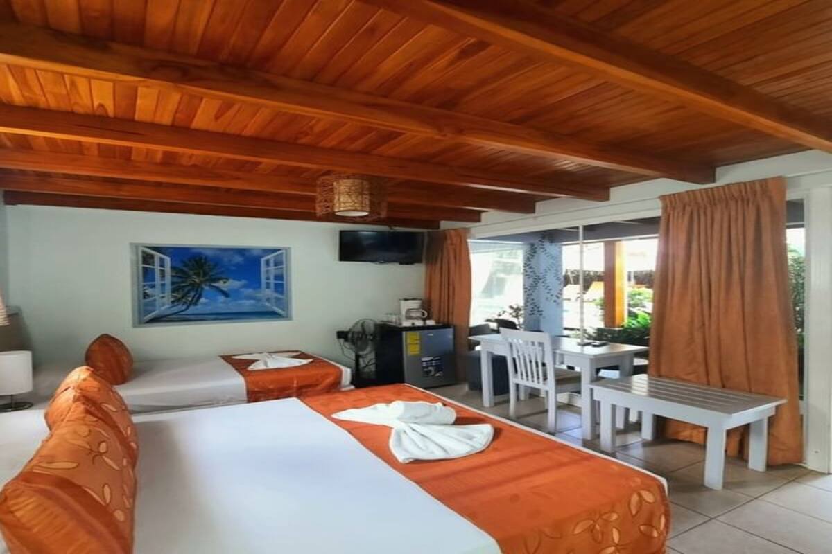 Una habitación de hotel con techo de madera, dos camas con ropa de cama naranja, una zona de comedor con mesa y sillas, un televisor montado en la pared y una ventana con cortinas.