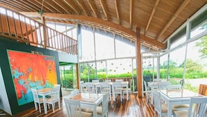 Un comedor amplio con mesas y sillas blancas, ventanas grandes que dan a zonas verdes y techo de madera.
